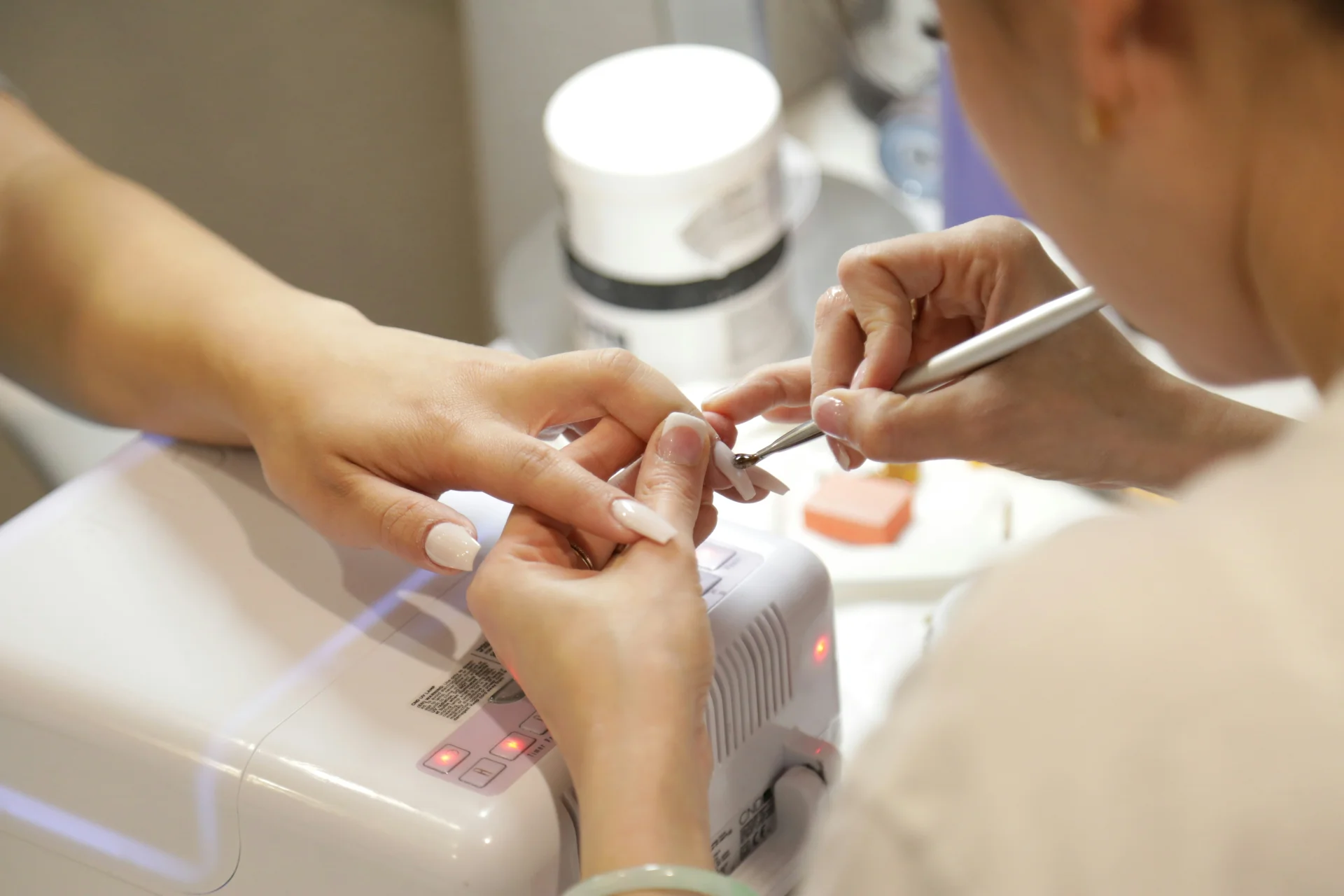 Kobieta siedzi w salonie kosmetycznym, gdzie stylista wykonuje jej manicure hybrydowy.
