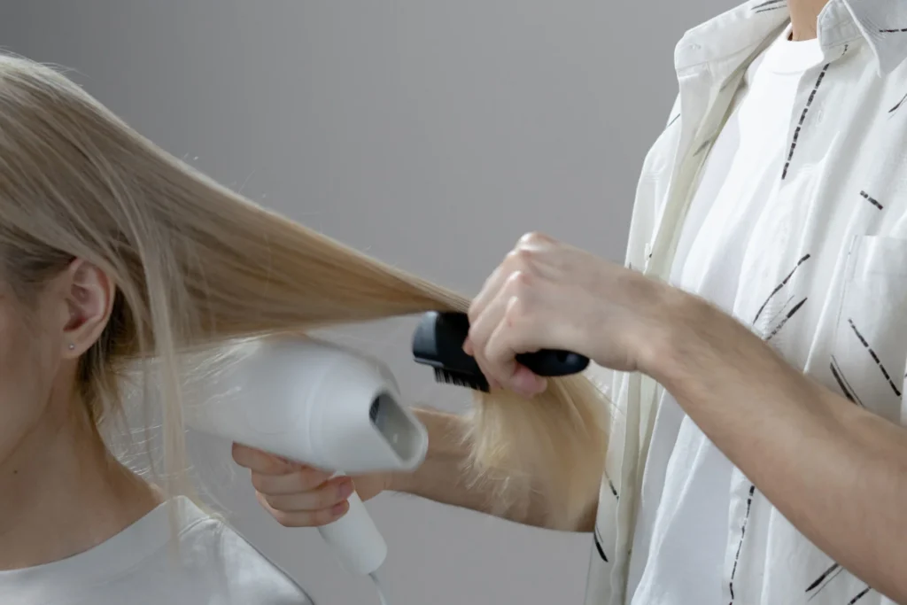Fryzjerka używa suszarki i szczotki do stylizacji długich, blond włosów klientki w salonie.